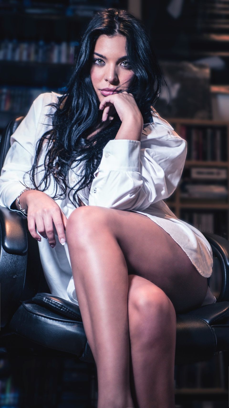 A mature woman sitting in a leather chair wearing nothing but an oversized white shirt.